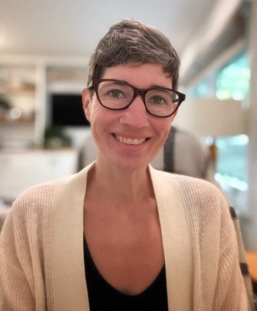 A headshot of Betsy who is smiling at the camera. They are wearing dark rim glasses, a white seater and a dark shirt.