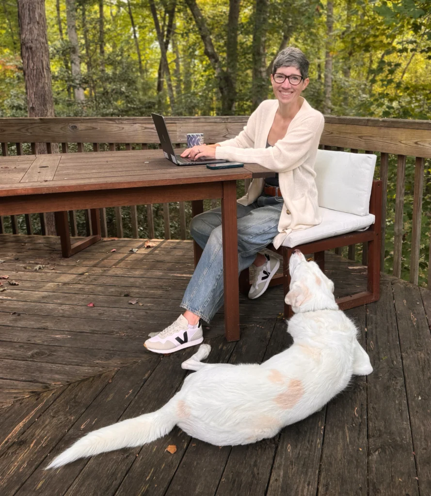 Betsy sitting as a table looking up from her laptop and smiling at the camera. A large white dog is at her feet looking up at her. The deck is surrounded by green, leafy trees.