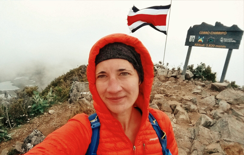 Betsy, wearing an orange puffy coat, stands on the top of Cerro Chirripó. A Costa Rican flag flies in the wind behind her next to a sign marking the summit. It's foggy but you can just make out a valley lake in the distance.