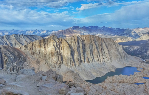 A view of a rocky ridge in the Sierra Nevada Mountains towering above an alpine lake. Mountains line the skyline in the distance beneath and blue sky with white clouds.