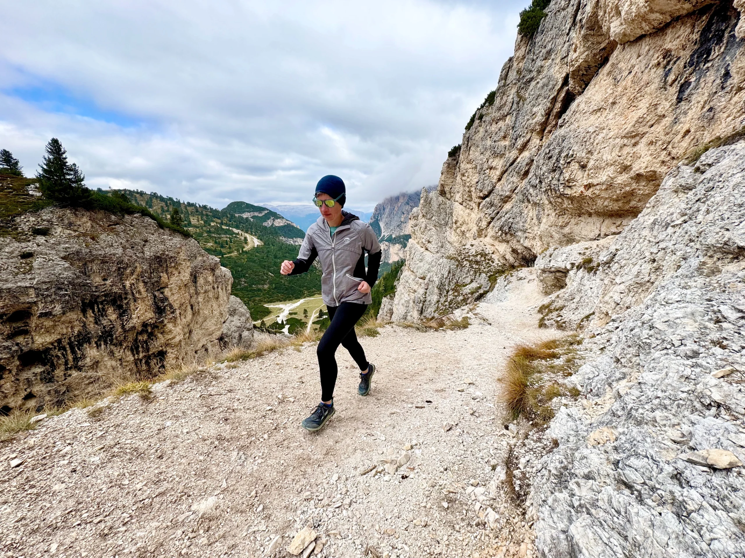 Betsy running up a rocky path in the Dolomites. A drop off on the left of the path reveals green valleys in the distance. Steep rocky cliffs line the right side of the path.