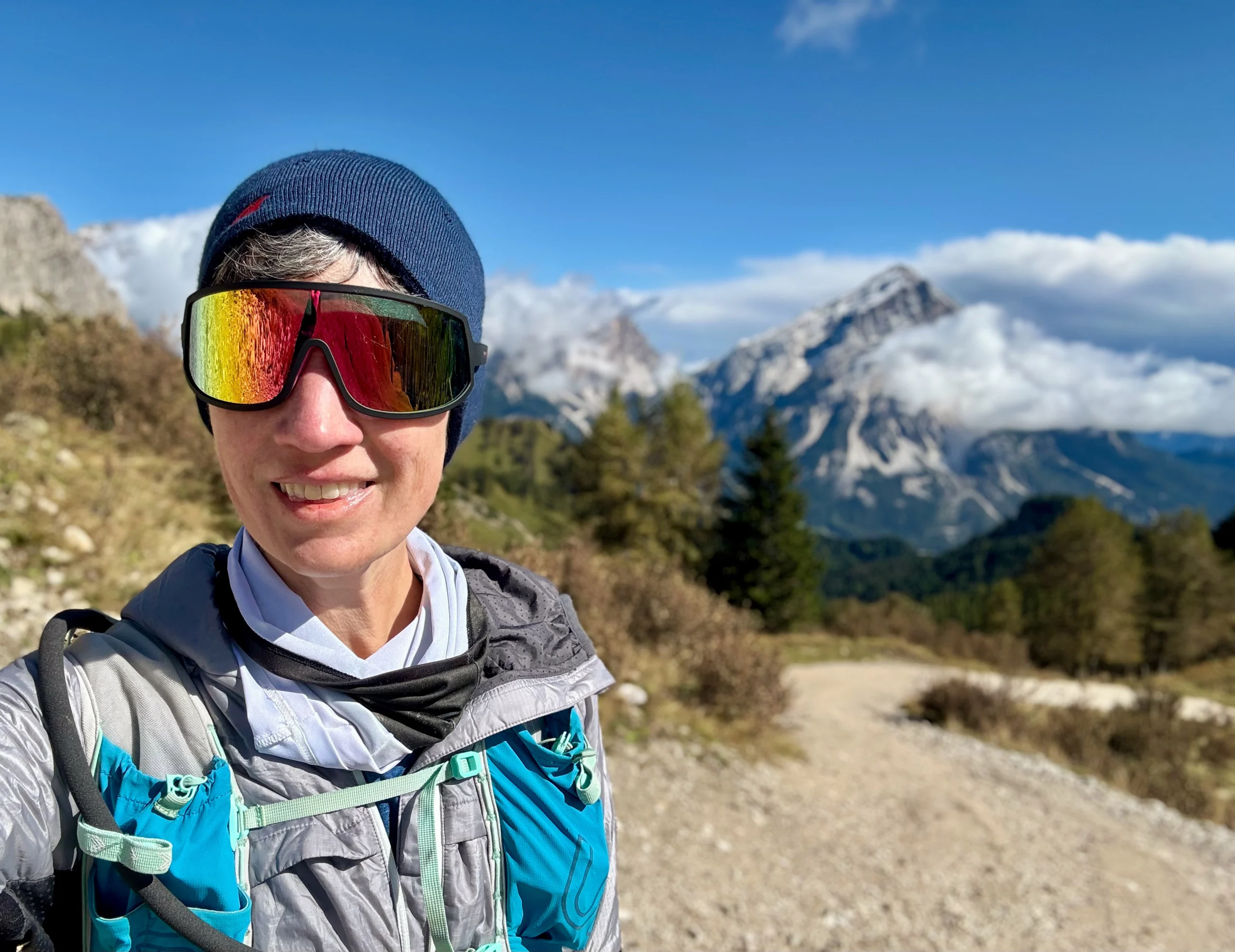 Betsy smiling at the camera. The background shows rocky mountains, green trees and a gravel path.