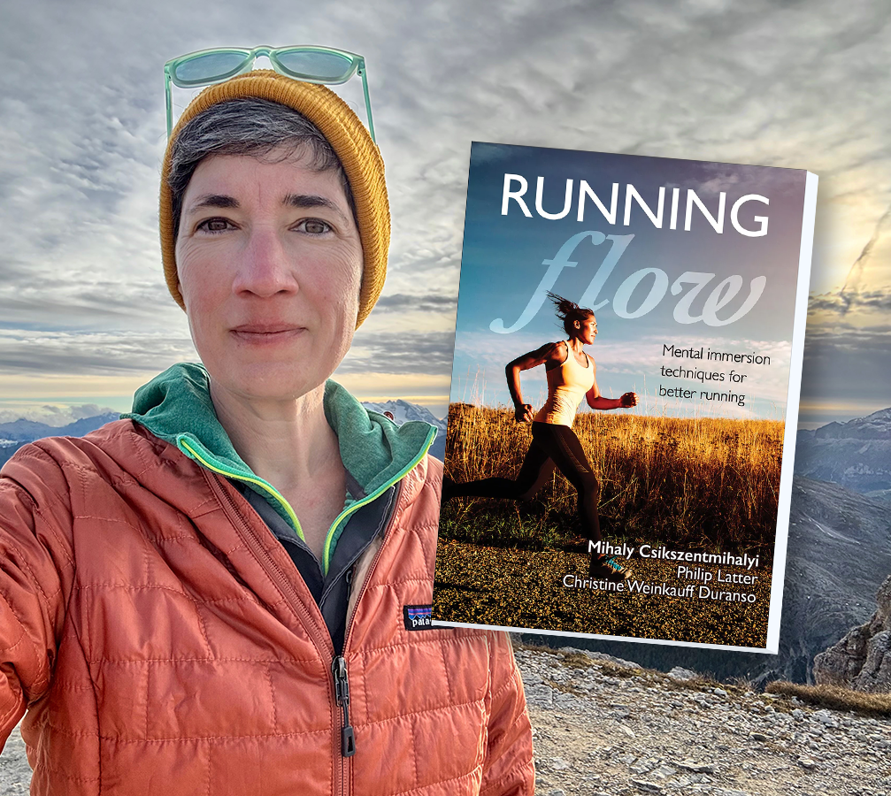 A selfie of Betsy looking at the camera, wearing a maple red jacket over a green shirt and mustard yellow beanie. She's standing in front of rocky mountains at sunset. An image of the cover of the book "Running Flow" overlays the image to the right of Betsy.