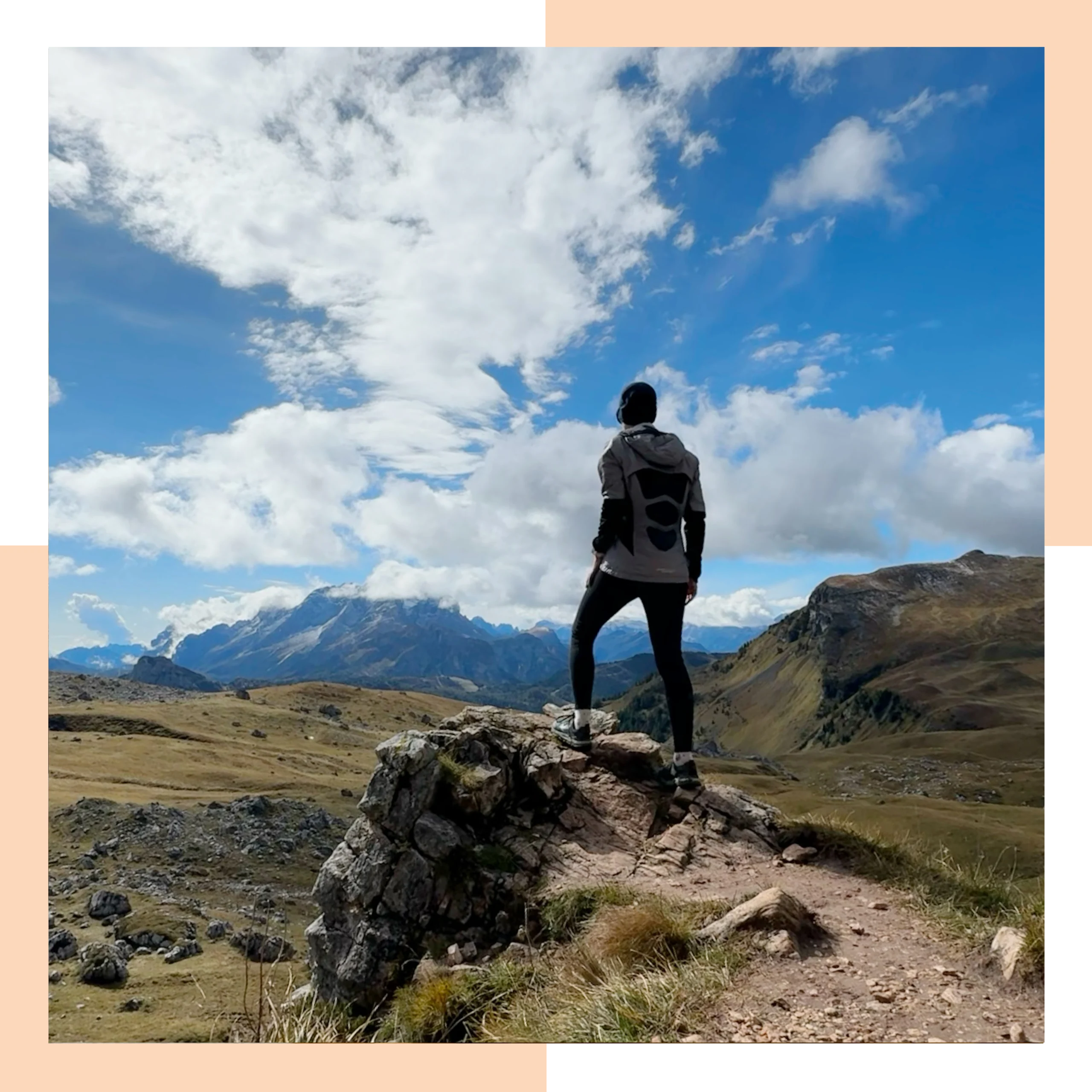 A runner standing on a rock looking out over a valley with rocky mountains in the distance. It's sunny with blue sky and fluffy white clouds.