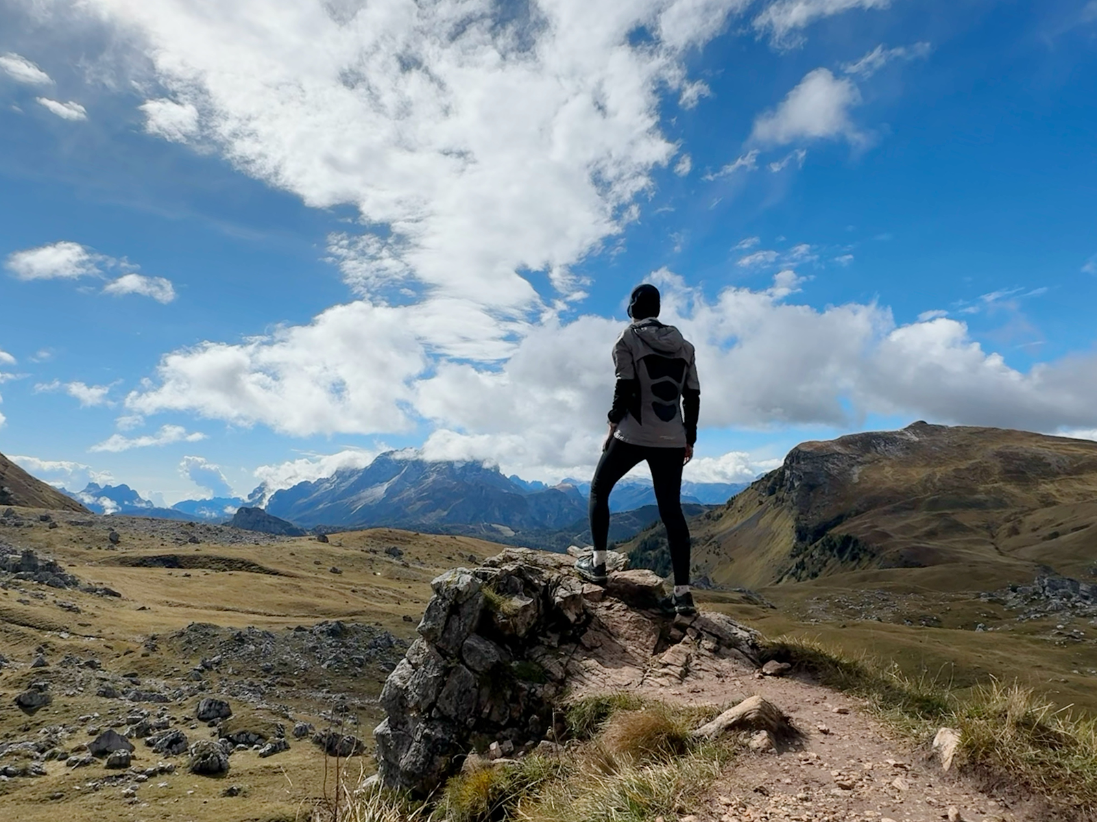 A solo runner stops to take in the view of a grass-covered plateau in front of distant rocky mountains beneath a blue sly with puffy white clouds.